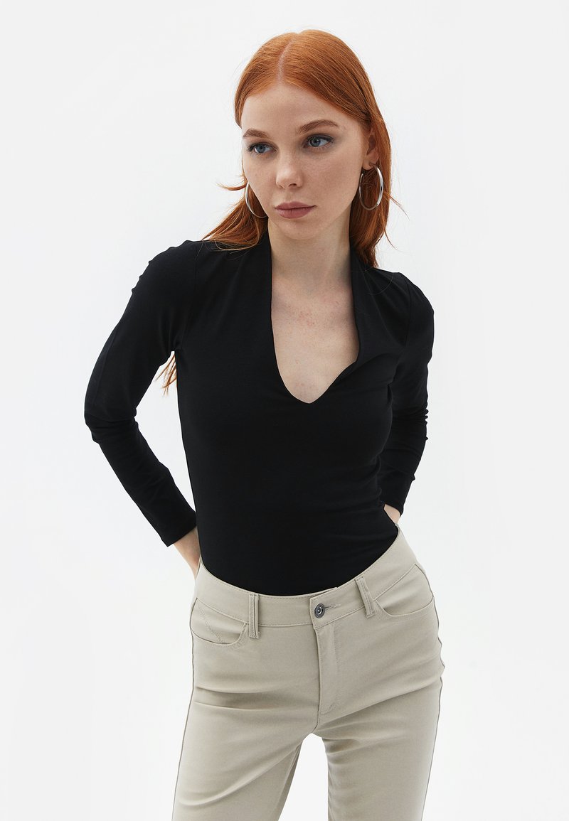 Langærmet sort bodystocking med dyb V-udskæring og tætsiddende silhuet, parret med lyse beige bukser; blødt stof og minimal syning.
