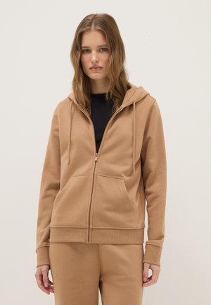 Junge Frau mit langen Haaren trägt beige Kapuzenjacke mit Reißverschluss und passende Hose, steht vor einem schlichten hellen Hintergrund.