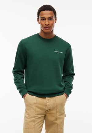Man wearing a dark green Tommy Jeans sweatshirt and beige pants with hands in pockets, standing against a plain white background.