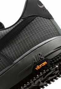 Zbliżenie na piętę czarnego buta sportowego z tkaniną w kratę, teksturowaną podeszwą oznaczoną napisem „AIR” i pomarańczowym logo „vibram” na bieżniku.