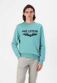 Tirkizna dukserica sa zaobljenim izrezom, dugim rukavima i grafičkim otiskom koji prikazuje "PME Legend American Classic" i akcentom krila.