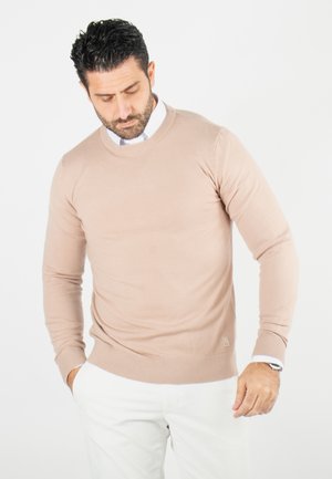 Pull beige à col rond en tissu doux, avec des manches longues et des poignets côtelés, associé à un pantalon blanc. Marquage subtil au niveau de l'ourlet.