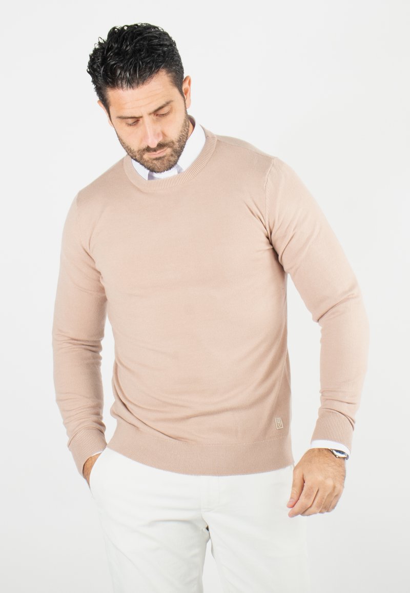 Pull beige à col rond en tissu doux, avec des manches longues et des poignets côtelés, associé à un pantalon blanc. Marquage subtil au niveau de l'ourlet.