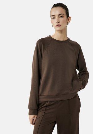 Kvinde med mørkt hår i en knold iført brun langærmet sweatshirt og matchende bukser, stående med den ene hånd i lommen foran ensfarvet baggrund.