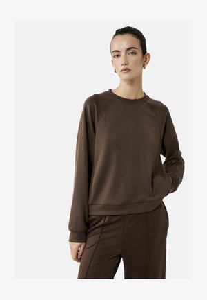 Kvinde med mørkt hår i en knold iført brun langærmet sweatshirt og matchende bukser, stående med den ene hånd i lommen foran ensfarvet baggrund.