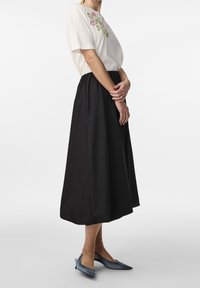 Frau in einem weißen Kurzarmoberteil mit pinkem Blumenstickerei, schwarzem A-Linien-Midikleid und blauen spitzen Slingbacks, die seitlich steht.