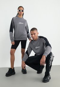 Grå hettegenser med svarte detaljer, med en logo. Svarte sykkelshorts. Svarte joggesko. To modeller poserer mot en ensfarget bakgrunn.