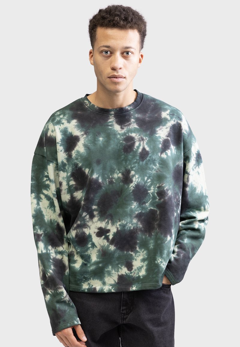 Ung mand med kort krøllet hår iført en grøn, sort og hvid tie-dye sweatshirt og mørke bukser, stående foran en ensfarvet baggrund.