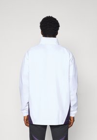 White jacket with a high collar, elastic cuffs, and purple side accents, featuring a smooth texture and loose fit. Simple style and design.