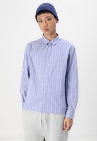 Light purple long-sleeve button-up shirt with white paint splatter and vertical stripes; paired with gray drawstring sweatpants and a navy beanie.