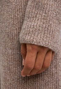 Maglione in morbidi toni beige con un motivo a costine testurizzato. Primissimo piano di una mano che afferra il polsino, evidenziando il materiale e la vestibilità.