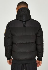 Sort dunjakke med hætte, der har et tekstureret, quiltet design og elastiske manchetter. Indeholder en synlig rem på bagsiden af hætten.