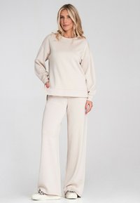 Beżowy sweatshirt z opadającymi ramionami i luźnym krojem, zestawiony z szerokimi beżowymi spodniami. Tkanina ma gładką, miękką fakturę.
