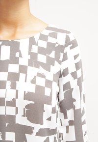 Pitkähihainen paita sileästä kankaasta, jossa on harmaa-valkoinen abstrakti neliökuvio ja pyöreä kaula-aukko.