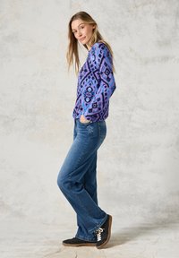 Paarse lange mouw top met geometrisch patroon, blauwe denim jeans en zwarte sneakers met witte veters. Model staat in zijprofiel.
