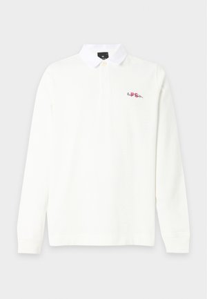 Joven de pie vistiendo una camiseta polo blanca de manga larga, pantalones anchos color marrón claro y zapatillas blancas sobre un fondo liso.