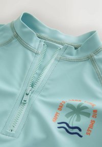 Un protector solar de color azul claro con un cuello alto, cierre de cremallera y un gráfico impreso de palmeras y olas con detalles en naranja y azul.