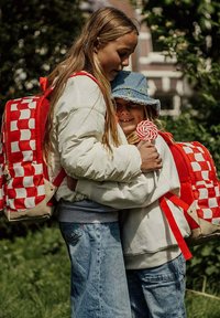 Rood-witte geruite rugzakken op twee kinderen buiten. De ene draagt een lichtblauwe denim hoed, terwijl de andere lang haar heeft en een witte jas draagt.