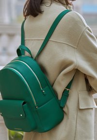 Grön läderryggsäck med rund design, som har två dragkedjefack och en framficka, bärs över en krämfärgad kappa.