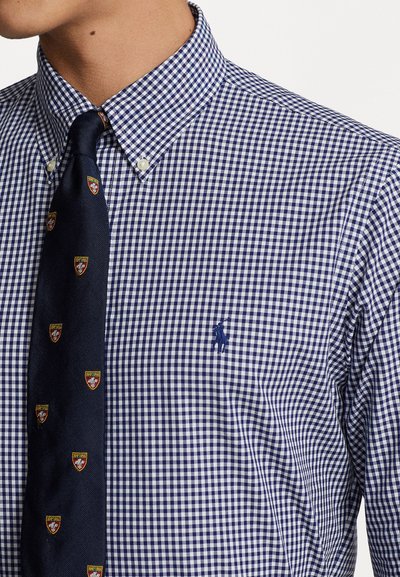 Chemise à carreaux bleu marine et blanc avec un col pointu et un logo de cheval ; assortie d'une cravate bleu marine ornée de petits emblemes.