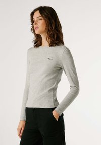 Camisa de manga longa cinza claro feita de tecido canelado. Apresenta um corte justo e um pequeno logótipo bordado no peito. Calças pretas mostradas.