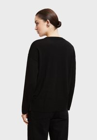 Maglione nero in maglia con vestibilità rilassata, dotato di un classico colletto girocollo e spalle basse. Indossato sopra pantaloni neri, che mettono in evidenza una consistenza morbida.