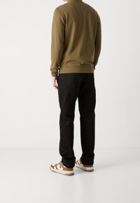 Man som bär en olivgrön tröja, svarta jeans och vit-bruna sneakers, står mot en enkel vit bakgrund med ryggen vänd.