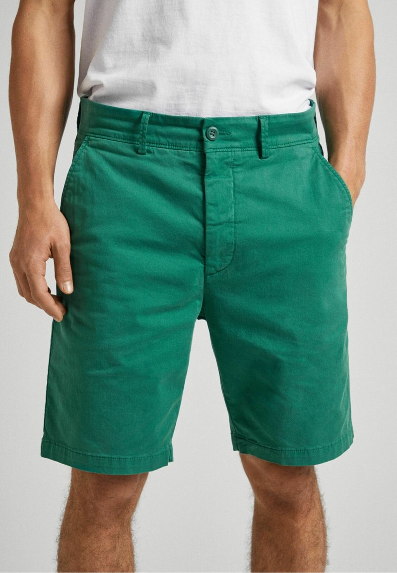 Homme portant un short vert jusqu'aux genoux et un t-shirt blanc, les mains dans les poches, debout devant un fond clair uni.