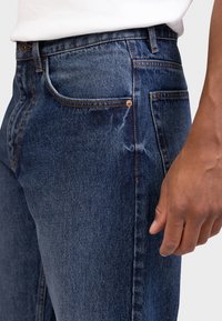 Σκούρο μπλε τζιν με ίσια γραμμή, με ορατές ραφές, μπροστινή τσέπη και λεπτομέρεια από χαλκό στην μέση.