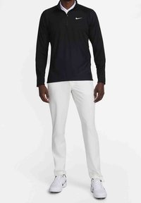 Polo nero a maniche lunghe Nike con zip a quarto, abbinato a pantaloni bianchi e sneakers bianche. Tessuto dalla texture liscia; logo sul petto.