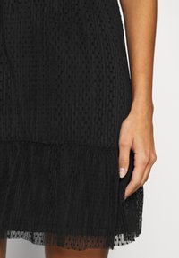 Jupe noire texturée avec un motif à pois et un ourlet à volants, montrée de la taille jusqu'à mi-cuisse, avec une main droite reposant doucement sur le côté.