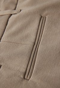 Beżowa tkanina o gładkiej strukturze, z dwoma pionowymi kieszeniami obszytymi ściegiem. Subtelny wzór tkania, neutralny odcień i starannie dopasowane elementy projektu.