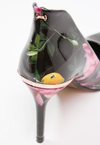 Chaussure à talon aiguilles noire avec motif floral brillant, bordure dorée à la base du talon, et petit noeud métallique à l'arrière.