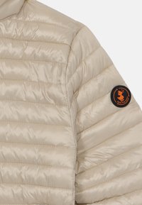 Beige insulated jacket featuring a quilted design, smooth texture, and a circular black logo patch with an orange duck emblem on the sleeve.