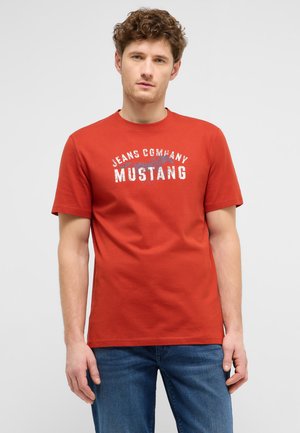 Junger Mann mit lockigem Haar, der ein oranges kurzärmeliges Shirt mit weißem "Jeans Company Mustang"-Schriftzug und blaue Jeans trägt.