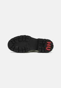 Svart gummisåle til sneakers med dype spor, som har et glatt sentralt område og rødt "HU"-logo på hælen. Teksturert for grep og holdbarhet.