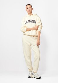 Egy nő áll egy krémszínű kapucnis felsőben és hozzá illő melegítőnadrágban, amin a "LAMANIA" felirat látható, fehér és szürke sportcipőt visel, semleges háttér előtt.