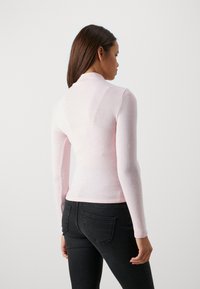 Maglietta rosa a maniche lunghe con collo alto, realizzata in un tessuto leggero e leggermente testurizzato. Indossata con jeans neri aderenti.