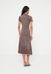 Žena s kučeravými vlasmi má na sebe hnedé zamatové midi šaty s krátkymi rukávmi a golierom, doplnené striebornými sandálmi na nízkom podpätku, stojaca s chrbtom otočeným k pozorovateľovi.