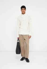 Suéter de punto crema con cuello alto y patrón de trenzas, pantalones beiges y una bolsa tote negra. Unos zapatos negros simples completan el conjunto.