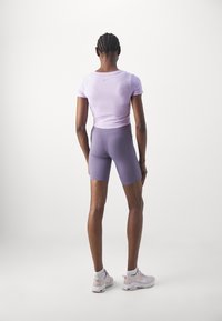 Person with braided hair wearing a light purple fitted short-sleeve top, matching cycling shorts, white socks, and pale pink sneakers, standing facing away.