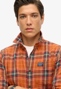 Camisa de franela a cuadros naranja con líneas contrastantes en azul y blanco, cuello con botones, bolsillo delantero con etiqueta de la marca y tejido texturizado.