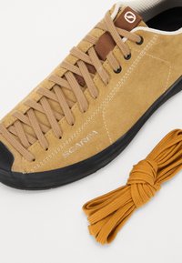 Tan suede shoe with black rubber sole and round toe, featuring light brown laces and a brown leather accent. Includes additional orange laces.