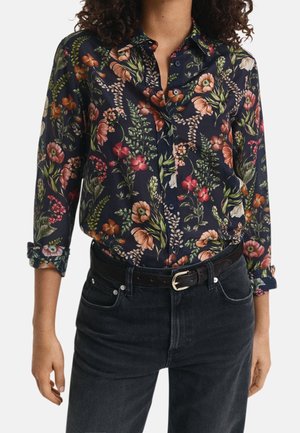 Kobieta w granatowej koszuli na guziki z kolorowym kwiatowym wzorem, z podwiniętymi rękawami, włożona do czarnych wysokich spodni dżinsowych z czarnym paskiem.