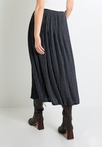 Falda midi plisada en tela gris oscuro, con una cinturilla alta, silueta fluida y acabado texturizado, combinada con botas de tacón marrones.