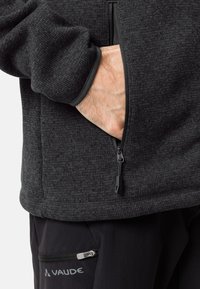 Veste en polaire noire texturée avec une poche zippée et des poignets élastiques, avec une main visible dans la poche. Logo Vaude sur le pantalon.