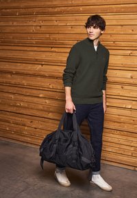 Sac de sport noir avec des bretelles réglables et plusieurs poches, tenu par une personne en pull vert foncé et jeans noirs, contre un mur en bois.