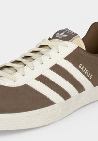 Καφέ σουέντ αθλητικό παπούτσι Adidas Gazelle με λευκές ρίγες, κρεμ κορδόνια και υφασμένο λευκό πάτο σε ανοιχτό φόντο.