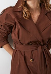 Hnědý trenčkot vyrobený z měkké tkaniny, s dvojitým zapínáním, širokými klopami, páskem v pase a ohrnutými rukávy s detaily na knoflících.