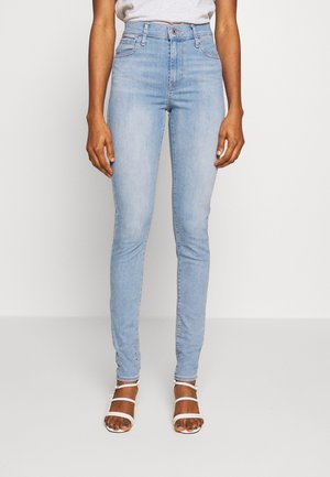 Personne portant un jean skinny bleu clair taille haute avec bouton et fermeture éclair, associé à des sandales blanches à talons et à brides, debout sur un sol blanc.